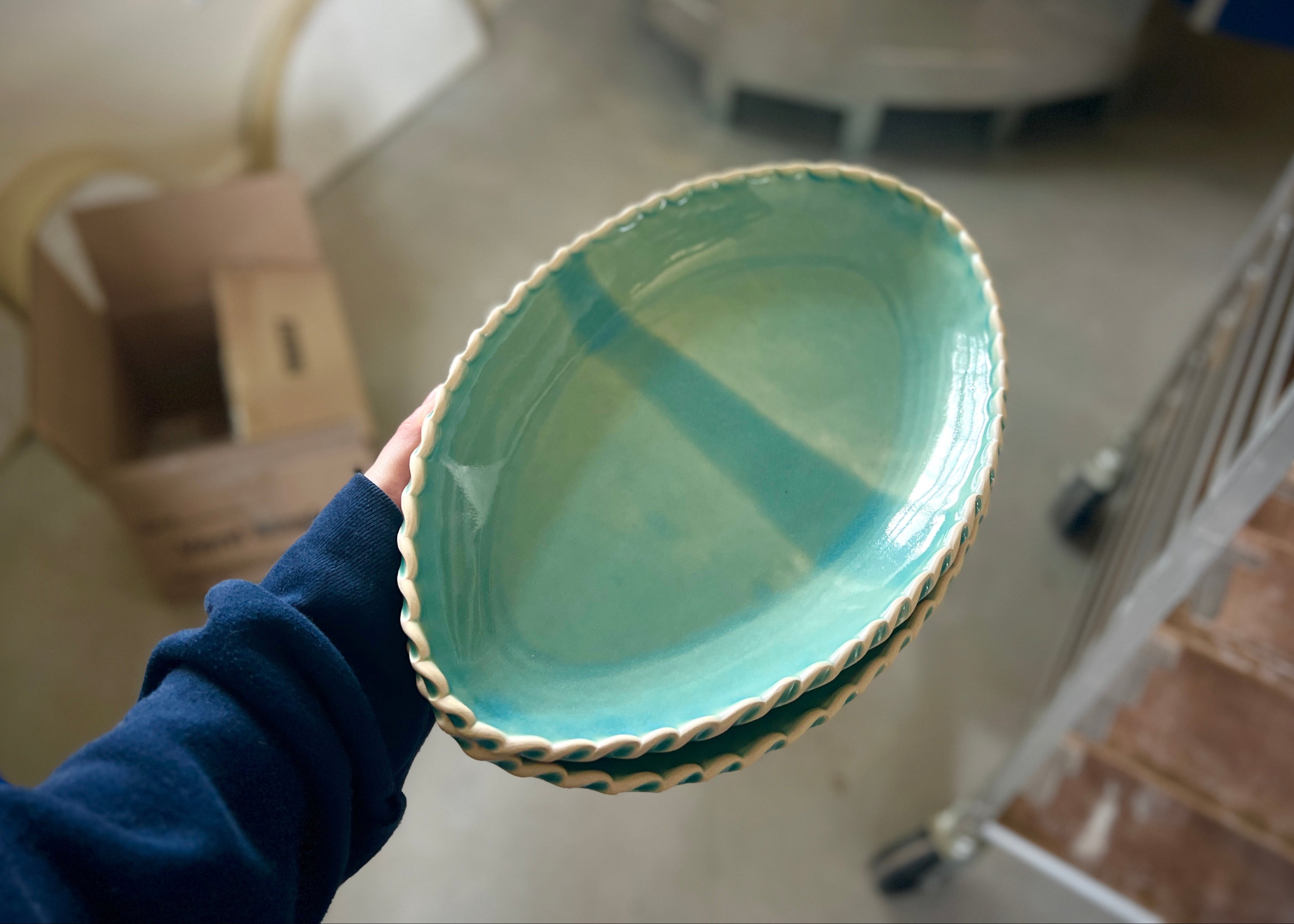 Person holding a teal ceramic bowl in an indoor setting with furniture and boxes in the background.